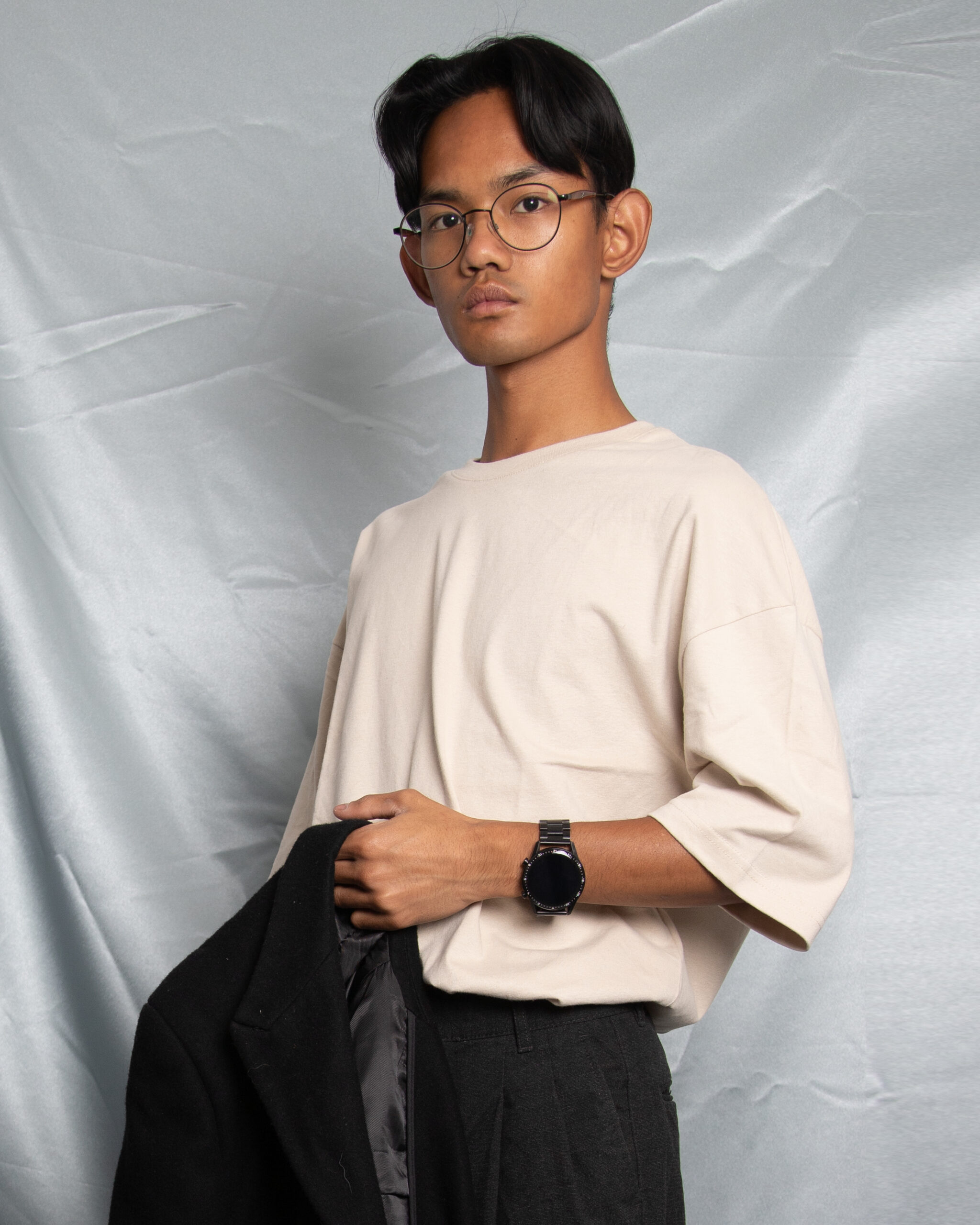 Upper body portrait photograph of of Chanphiphat Janthra, a young Thai man with glasses and short hair. He is against a satin sheet, holding his jacket.
