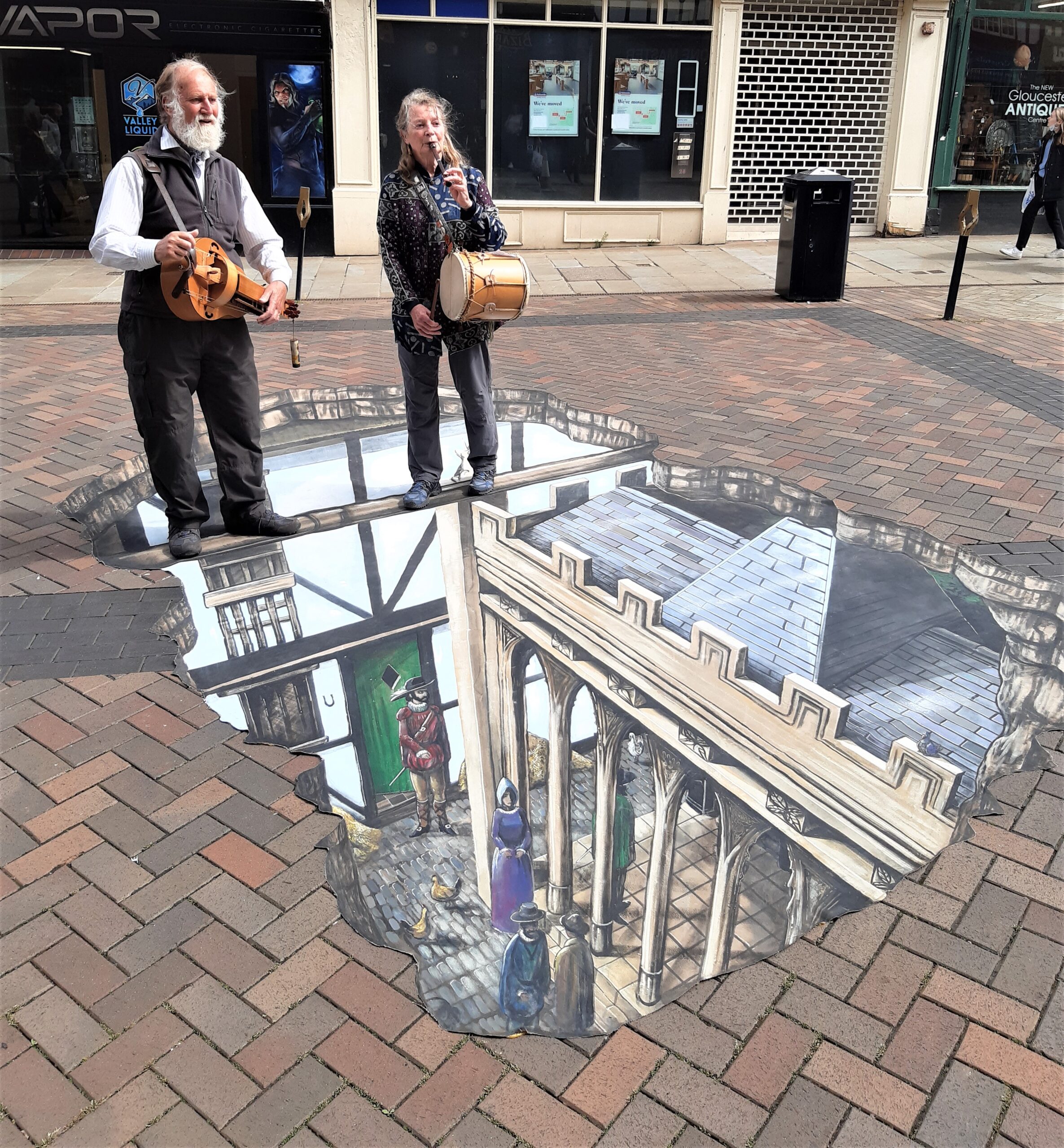 Photograph of two musicians in a public area, standing above mural of a mediaeval town - like a portal!
