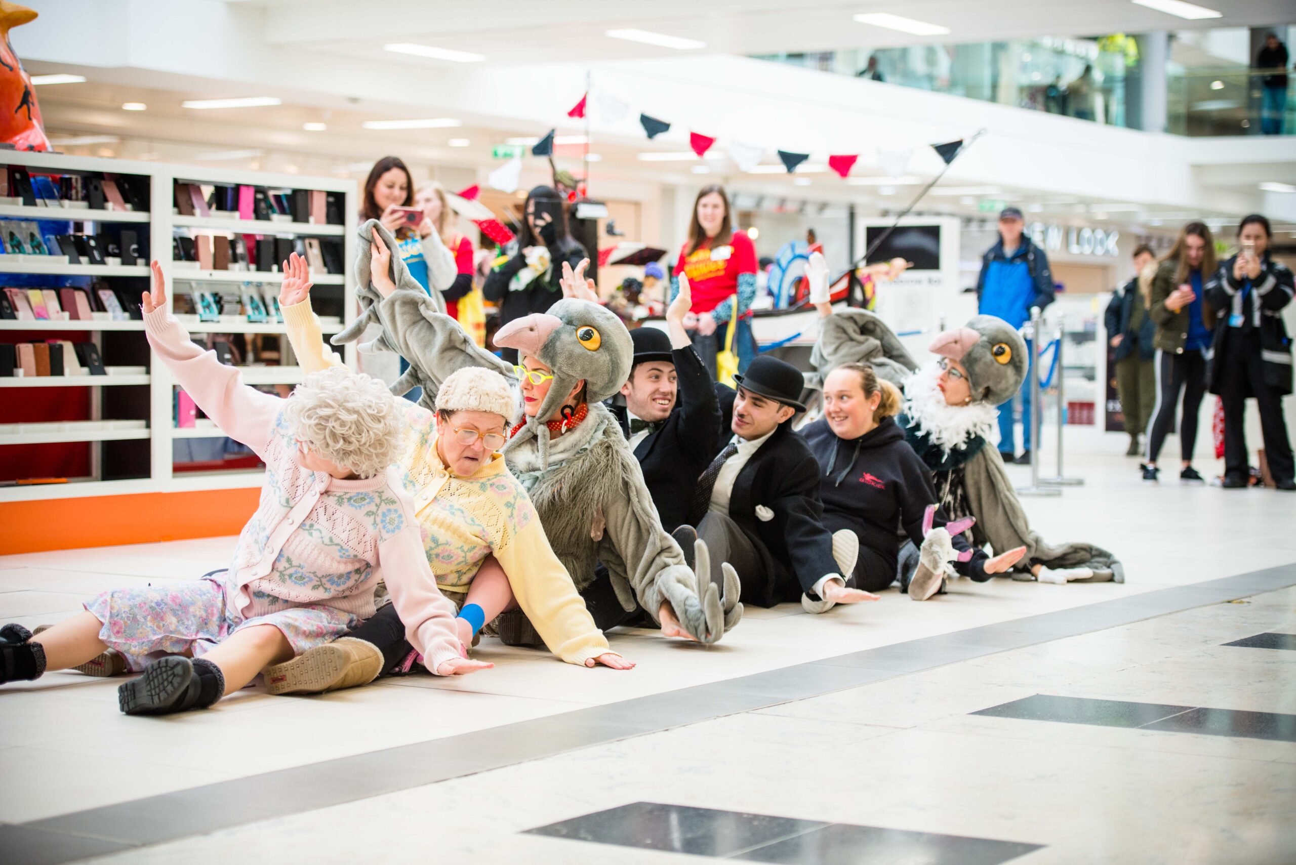 A photograph of group of people in a shopping centre. two of them wearing pigeon costumes and there are four people watching.