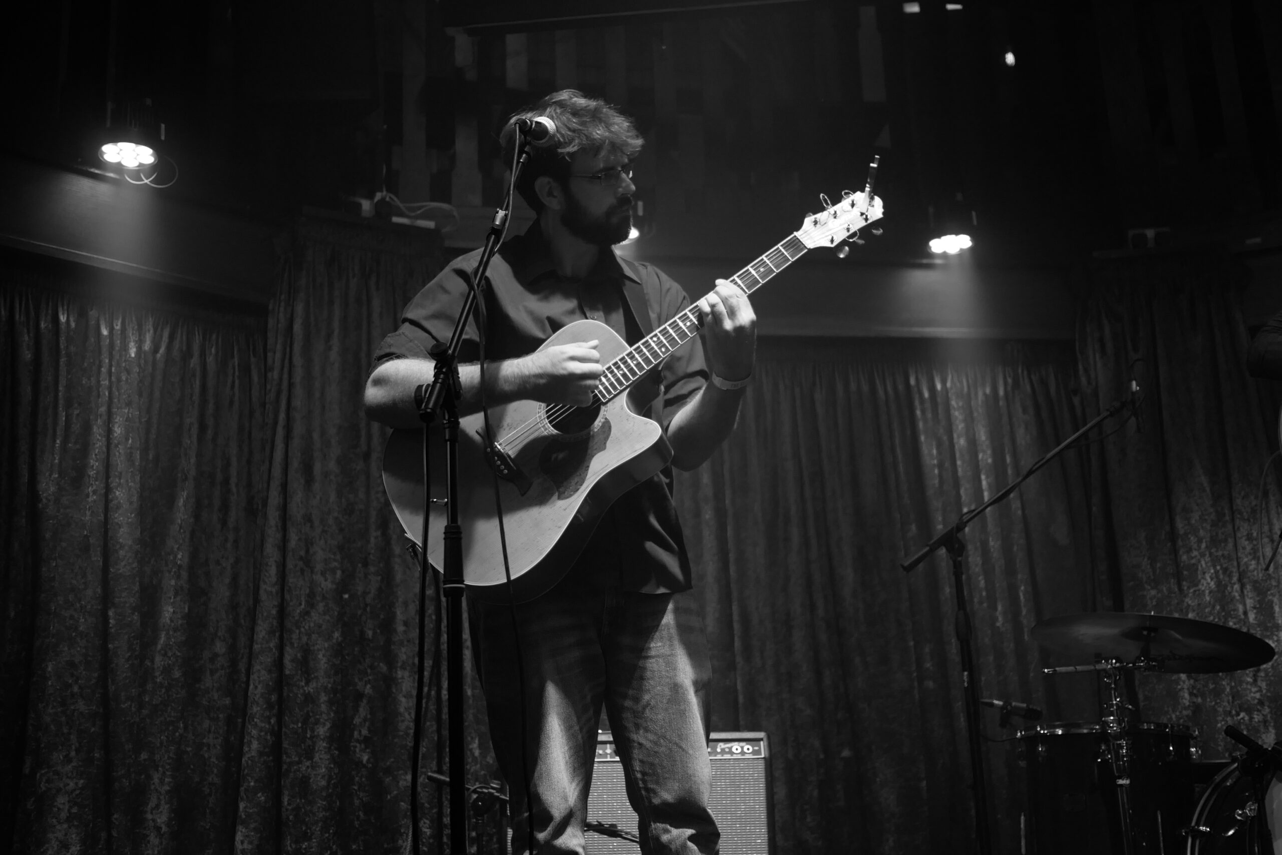 Photograph of a bearded man holding an acoustic guitar, staring at his guitar on stage.