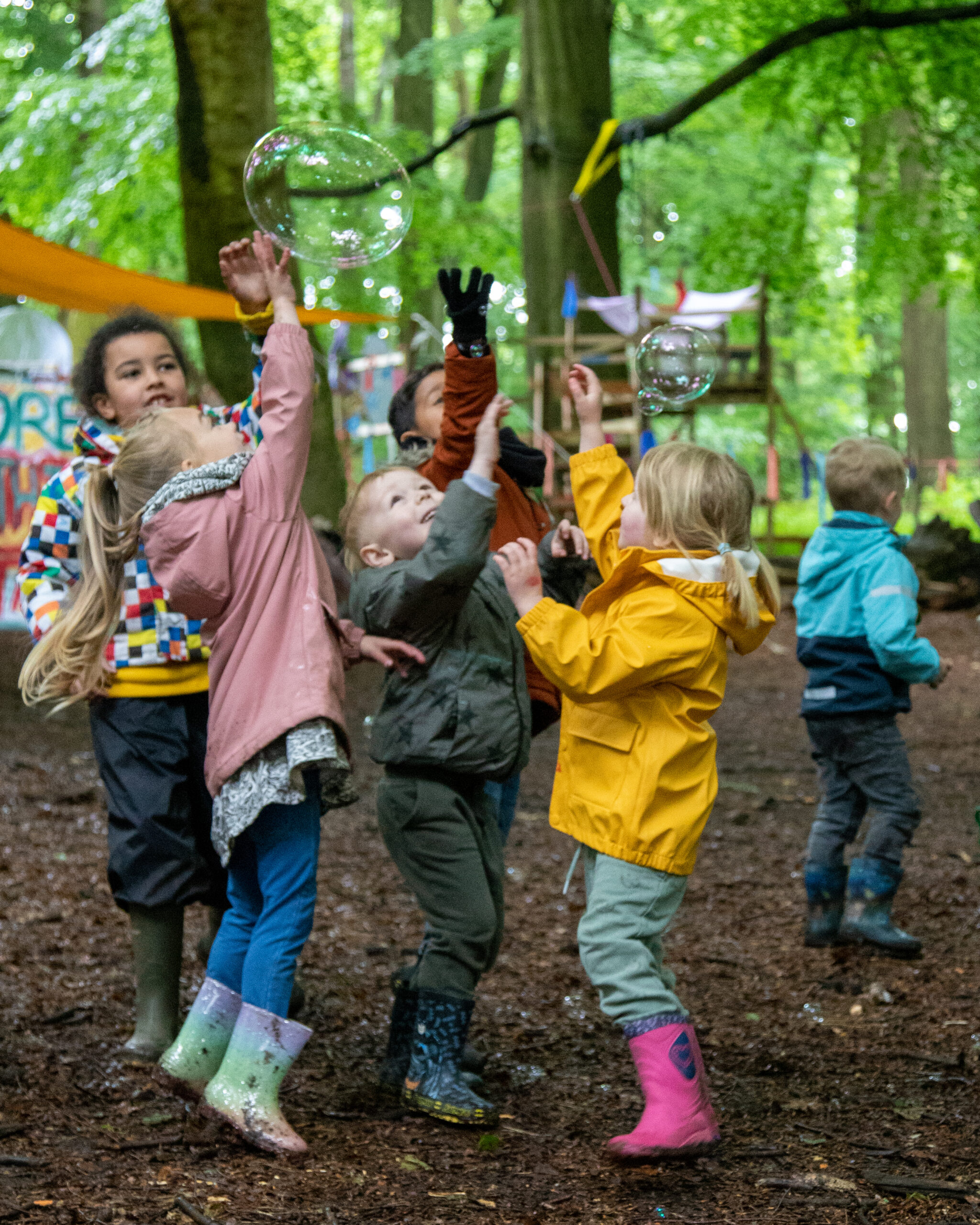 a Photograph of group of excited children playing with giant bubbles surrounded by trees.