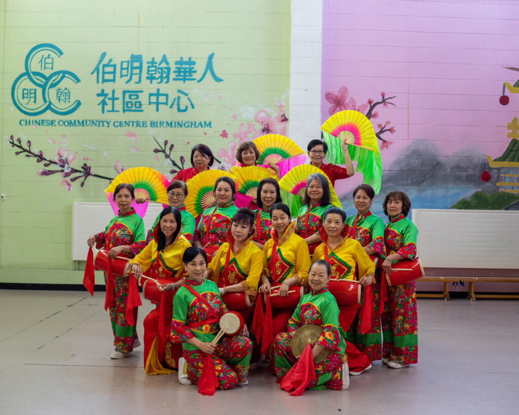 A photograph of group of women in traditional Chinese costume in front of a mural saying Chinese community centre Birmingham.