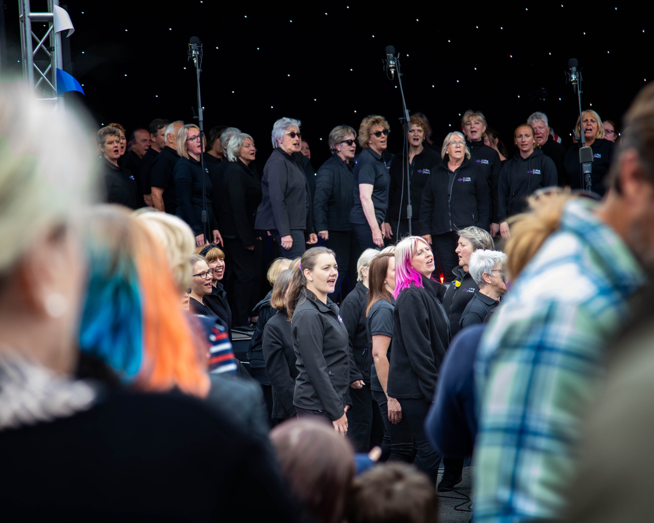 Photo of a choir singing on large stage - the crowd out of focus in front of them.