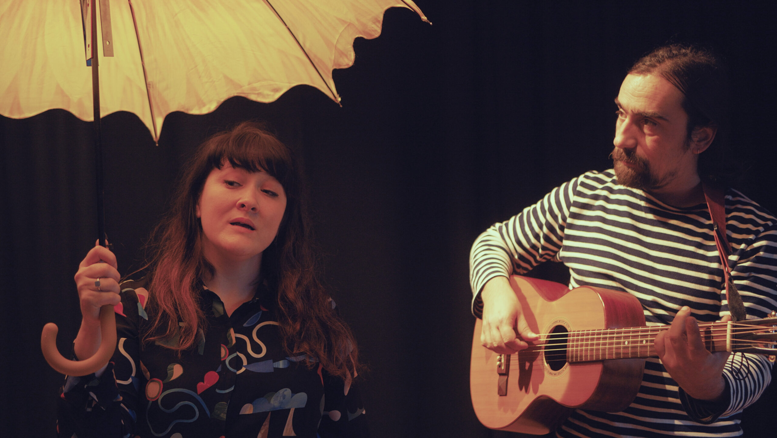 An image of two people. one person holding an umbrella and one person playing a guitar.