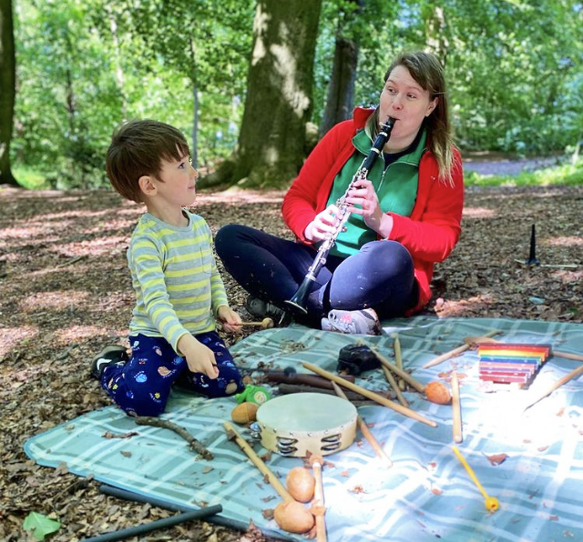 Photo of child in woodland, sat with a clarinet player at a picnic blanket.