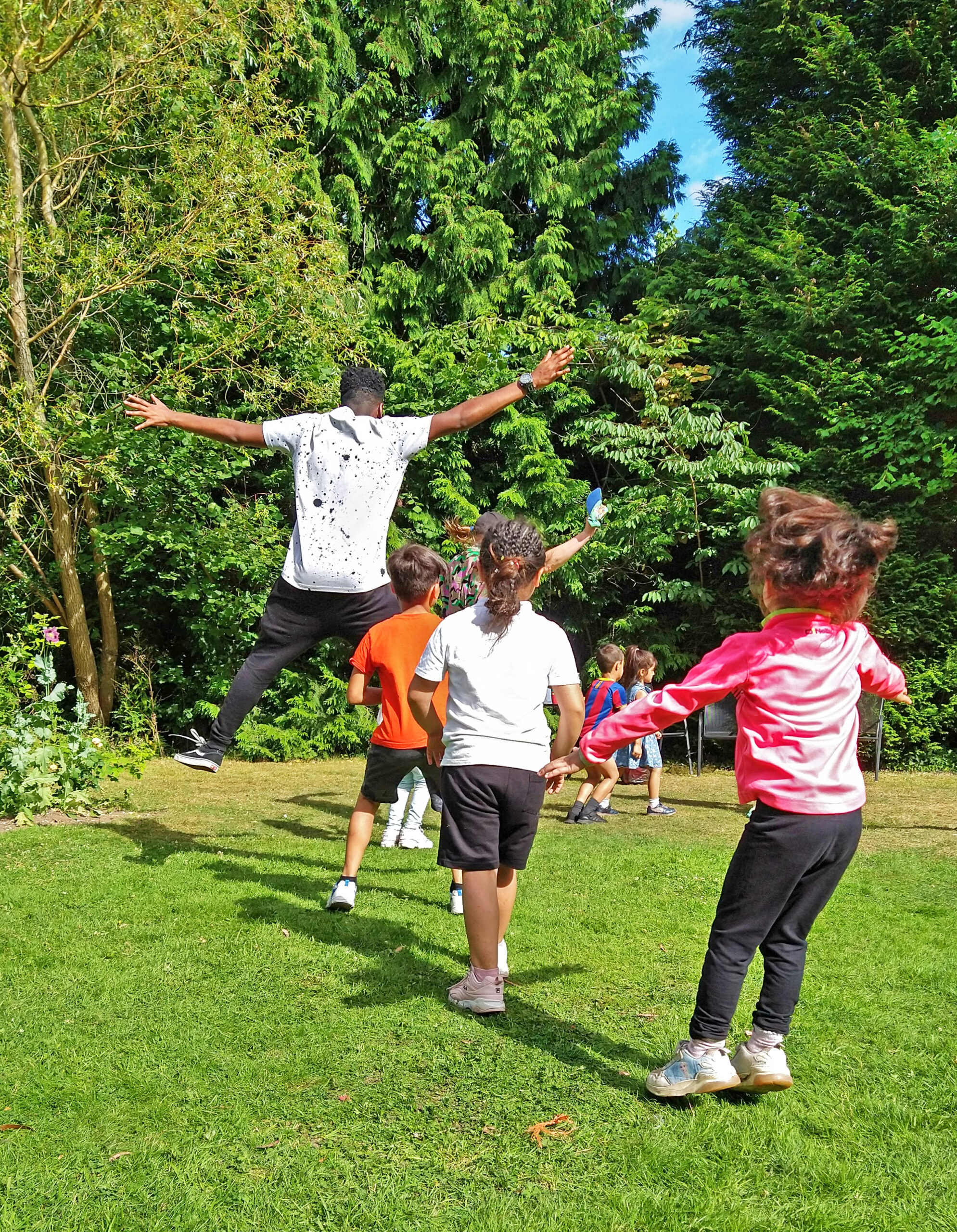 Photo of a line of children, running in a sunny park, with an adult jumping high, limbs outstretched