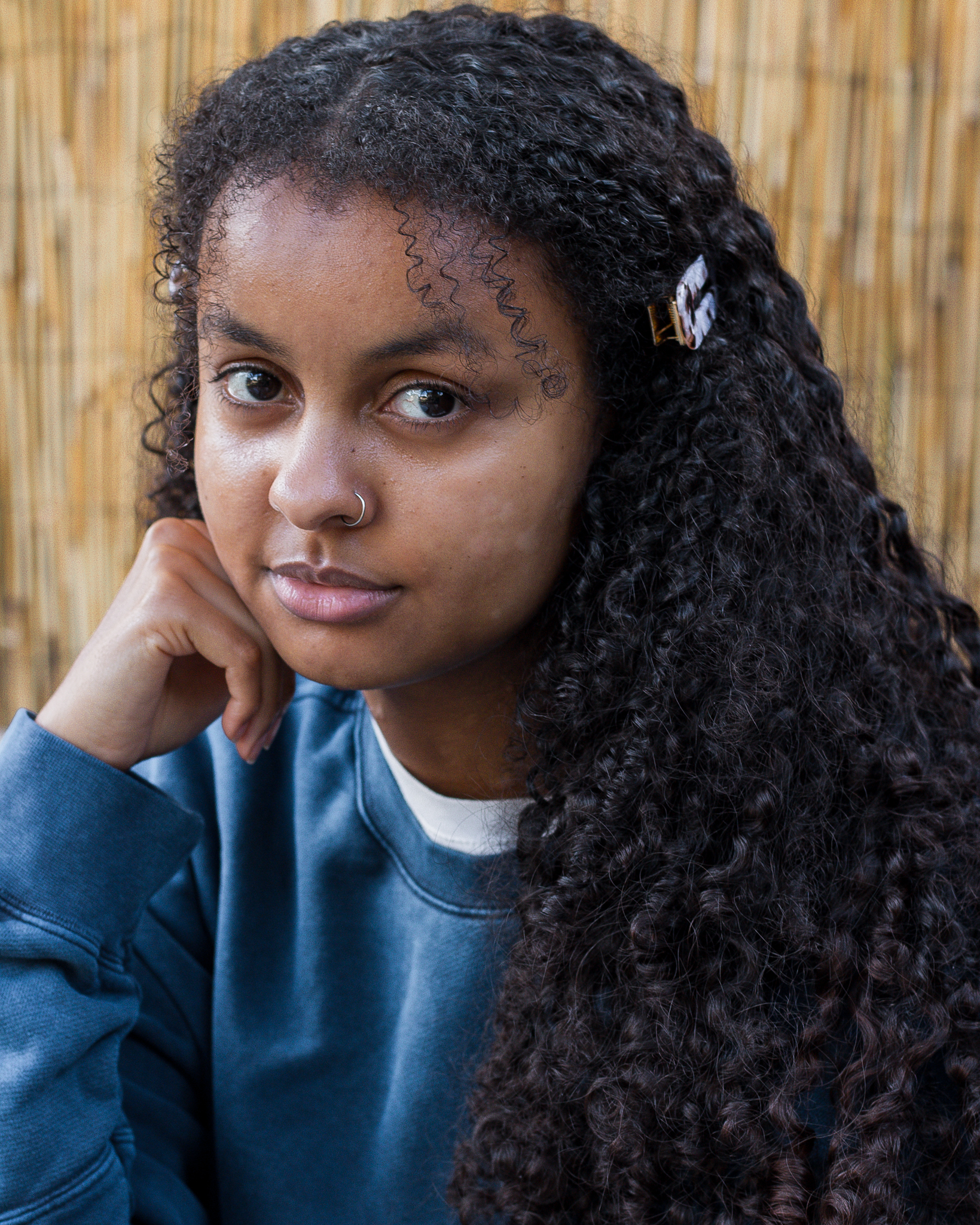 Headshot of Taneesha - a young black women with long, coiled hair, looking thoughtful with her head resting on her hand. She wears a blue jumper, and is in front of a straw background.