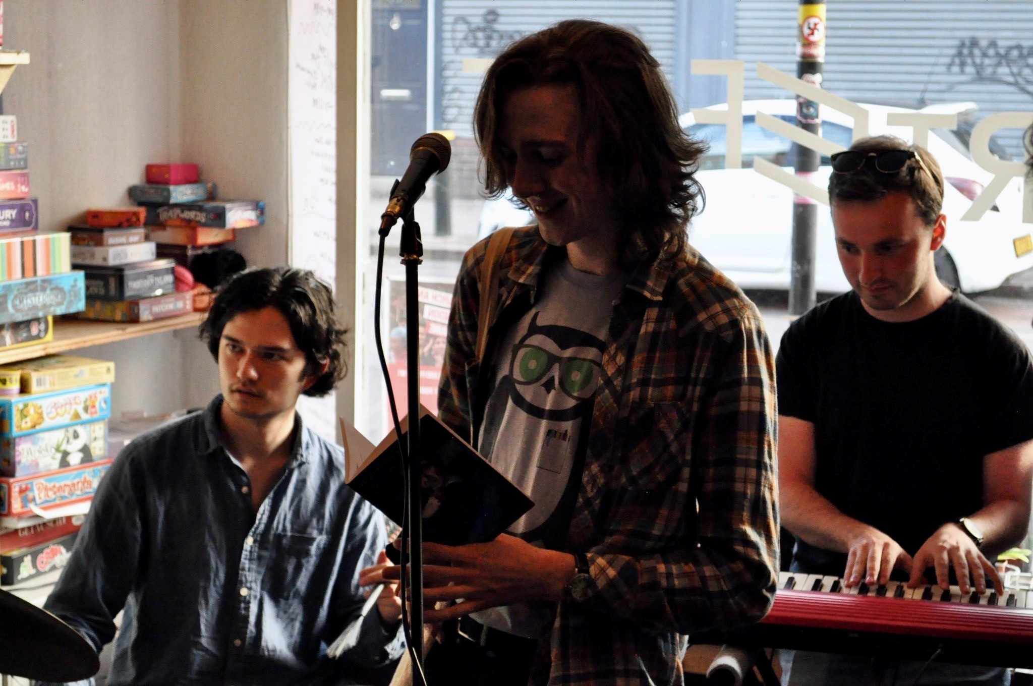 Photo of a white person, with medium-long hair, standing before a mircrophone and looking down at a pamphlet in their hand. They stand in front of a drummer, a keyboard player and a window.