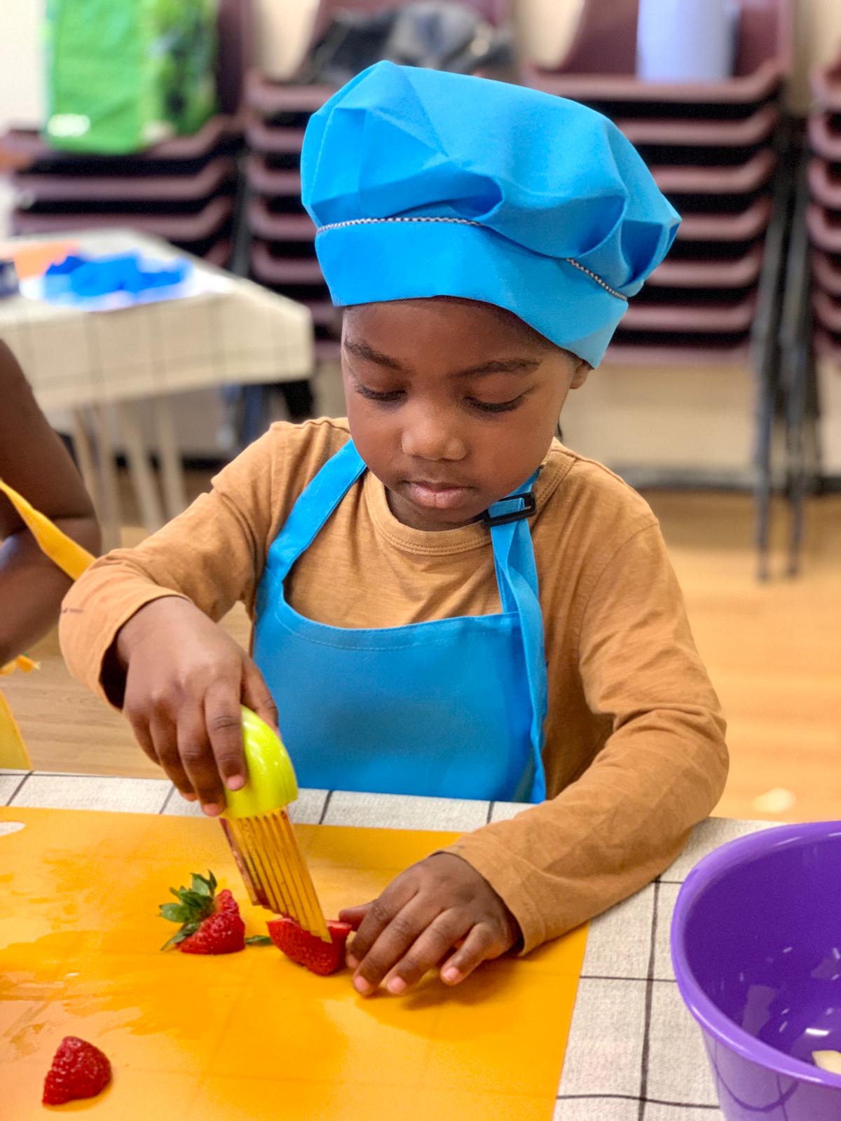 Photo of a very small child wearing a chefs hat and apron, cutting a strawberry
