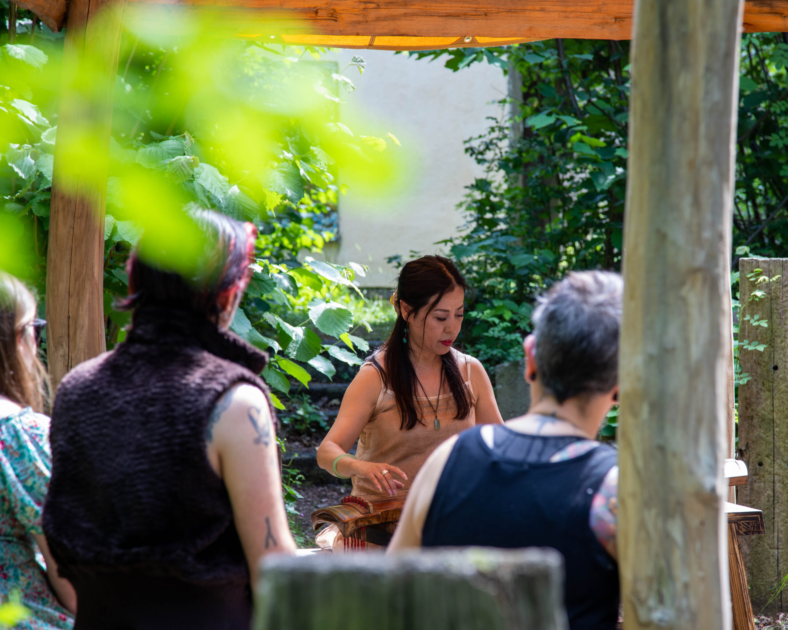 Photo of a woman playing the guqin, in a woodland sat down, with an audience and leaves obscuring the view.