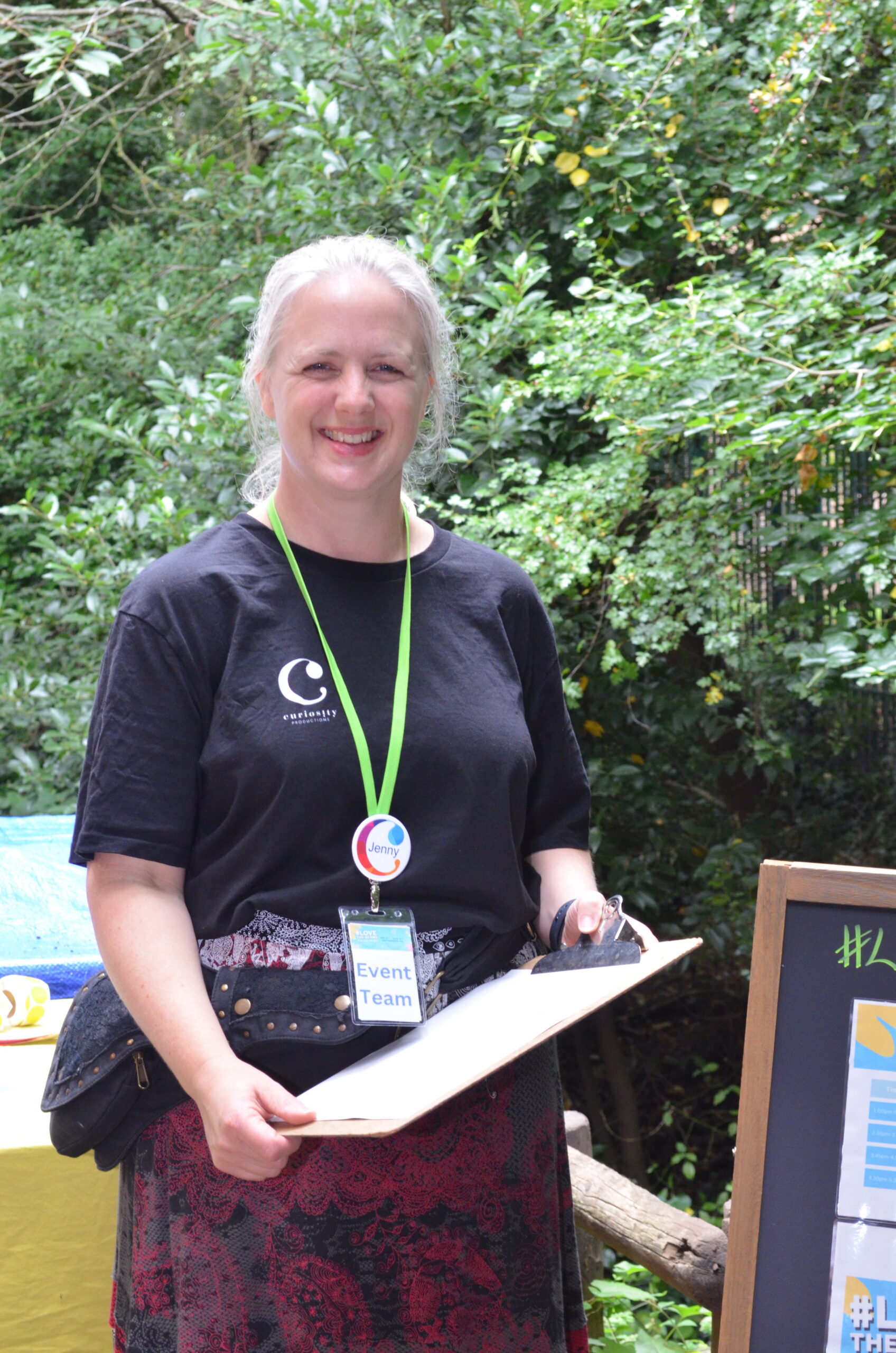 Jenny Martin, has blonde hair and fair skin. She is wearing a black t-shirt with the Curiosity Productions logo on, with a red and black long skirt and a green lanyard.