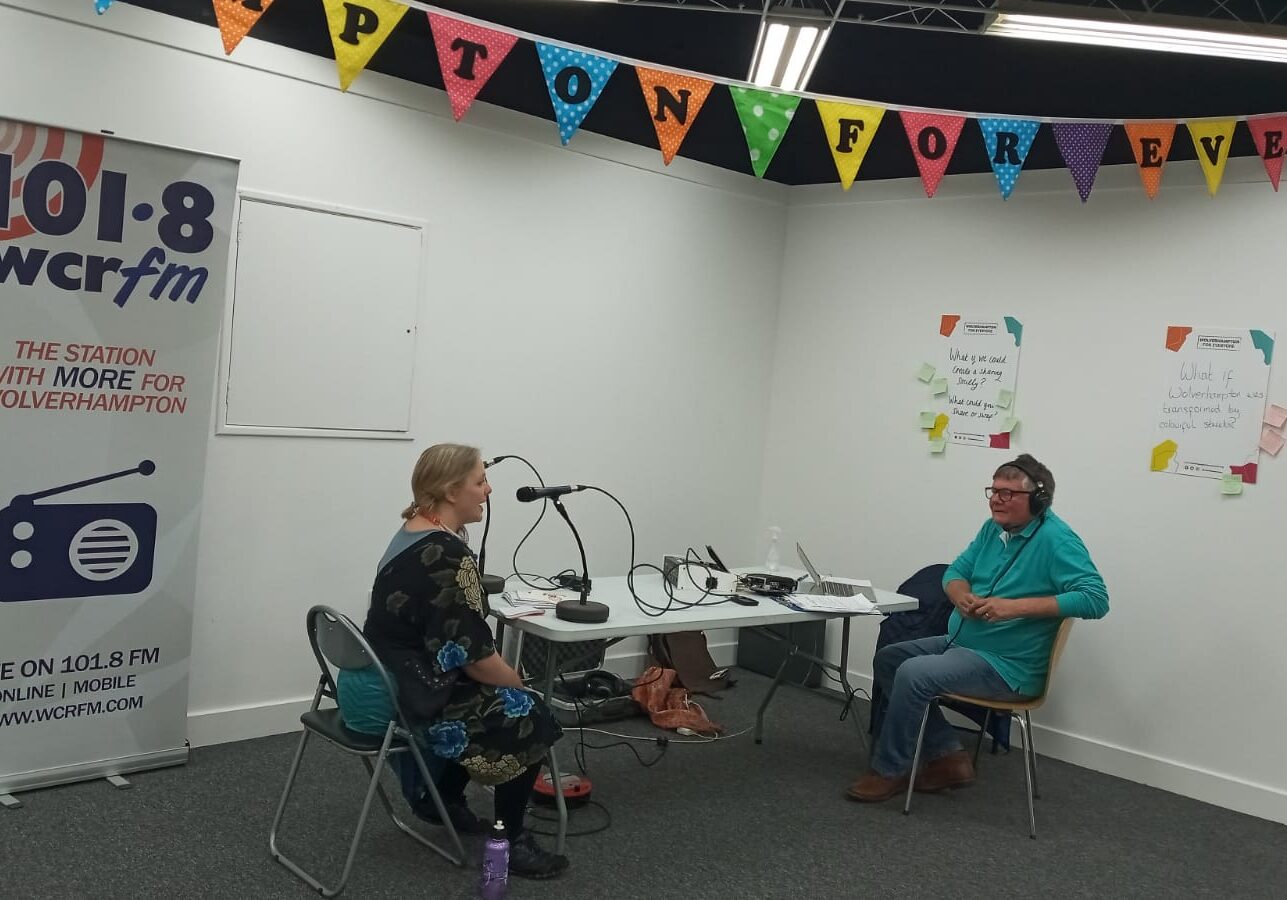 Photograph of Creative Director Jenny Smith being interviewed at a table in the corner of the room. To the left is a radio banner, reading WCR radio.